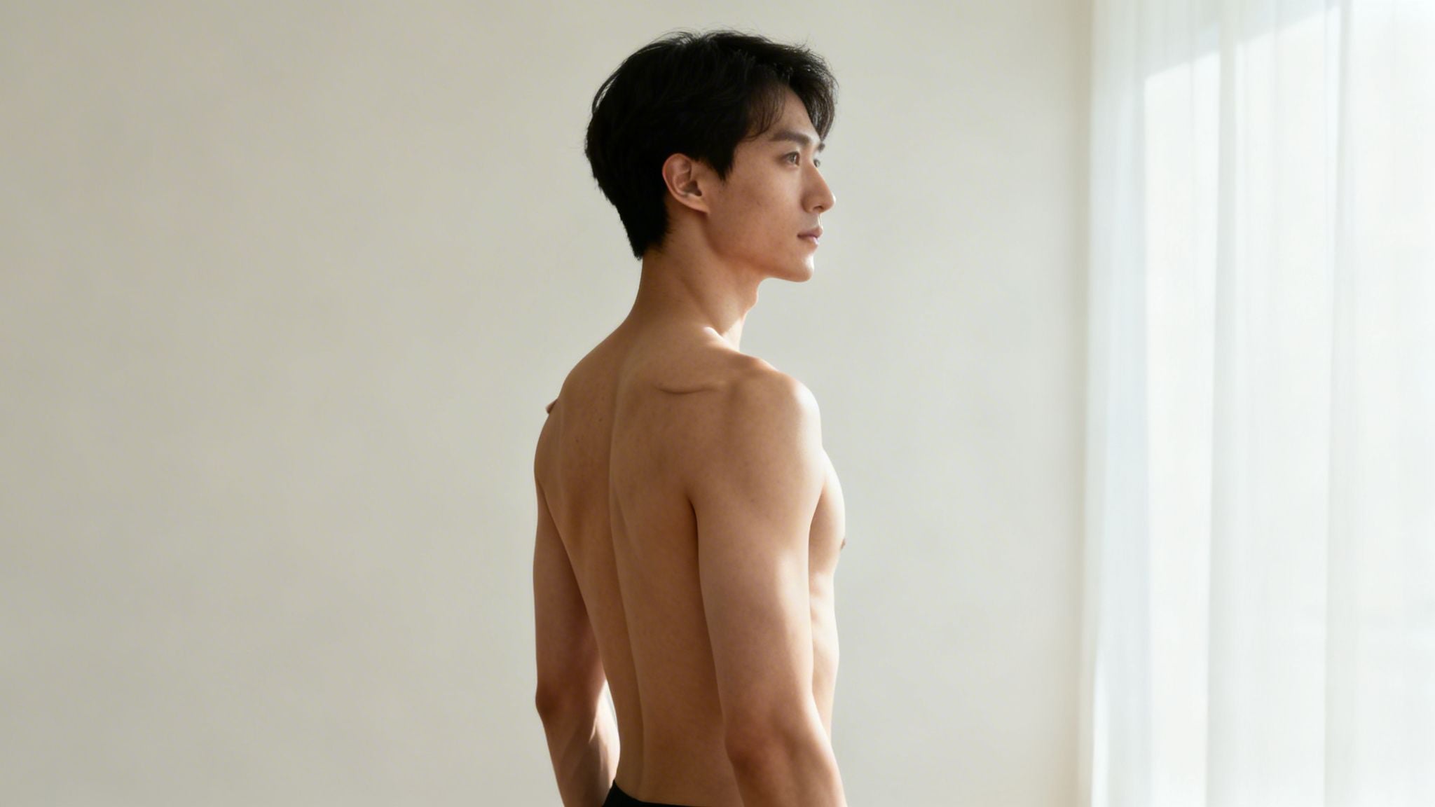 A shirtless young man with dark hair, seen from the side, looking right in a bright room.