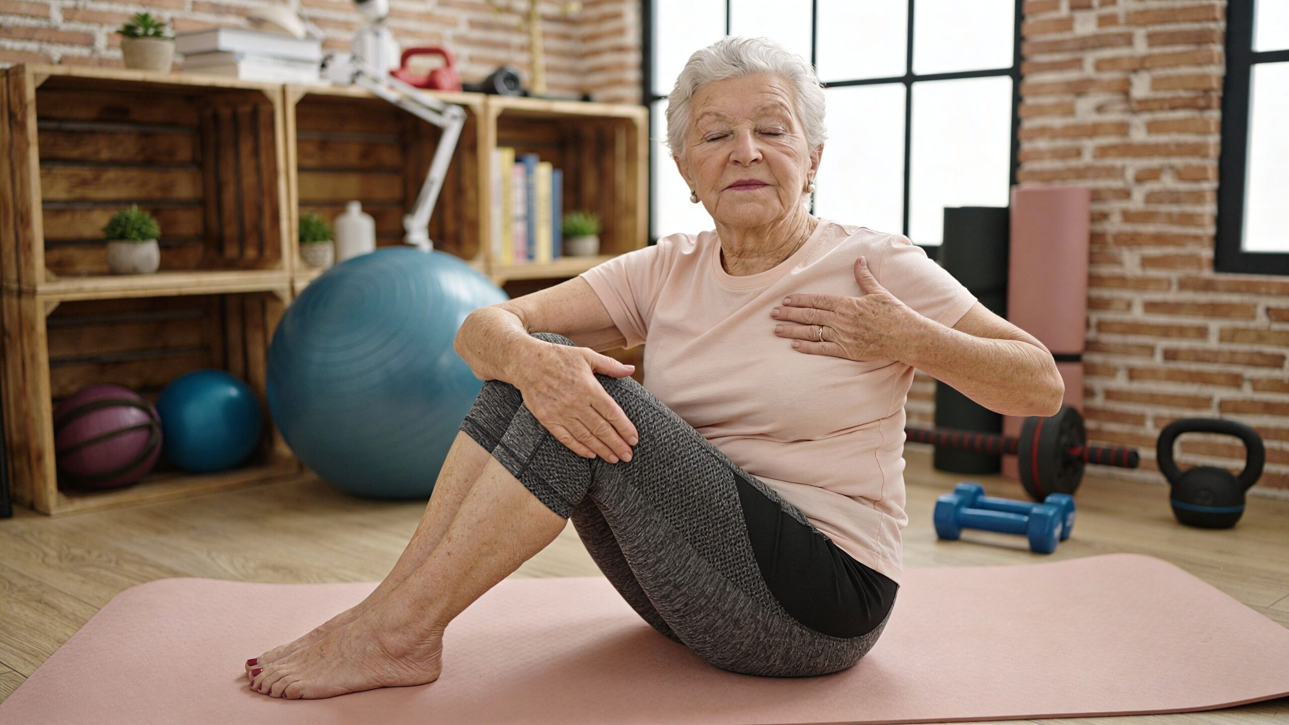 An elderly woman with grey hair practicing a seated dynamic chest stretch exercise on a yoga mat