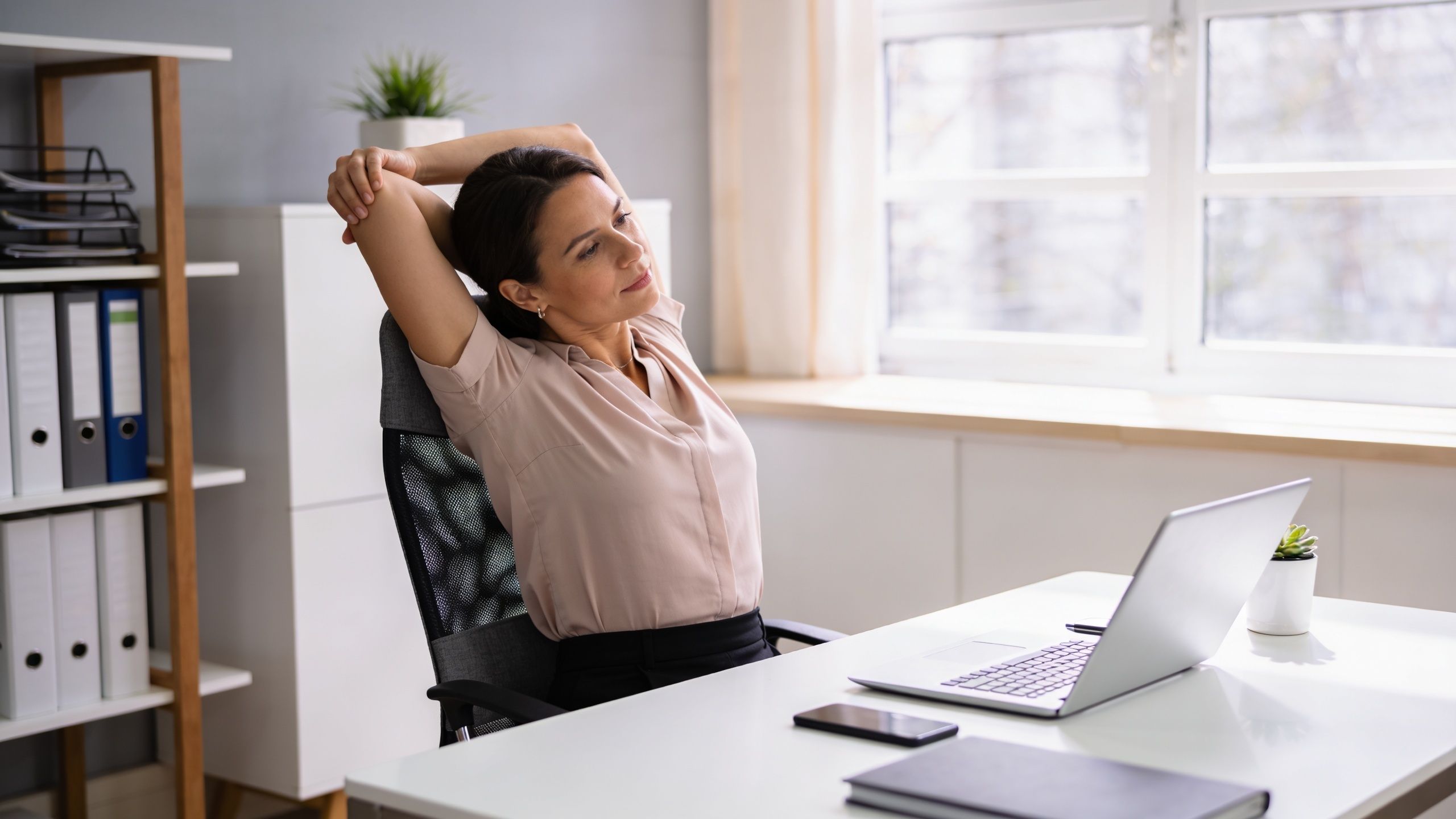 A professional woman stretching her arms behind her head while sitting at her office desk.