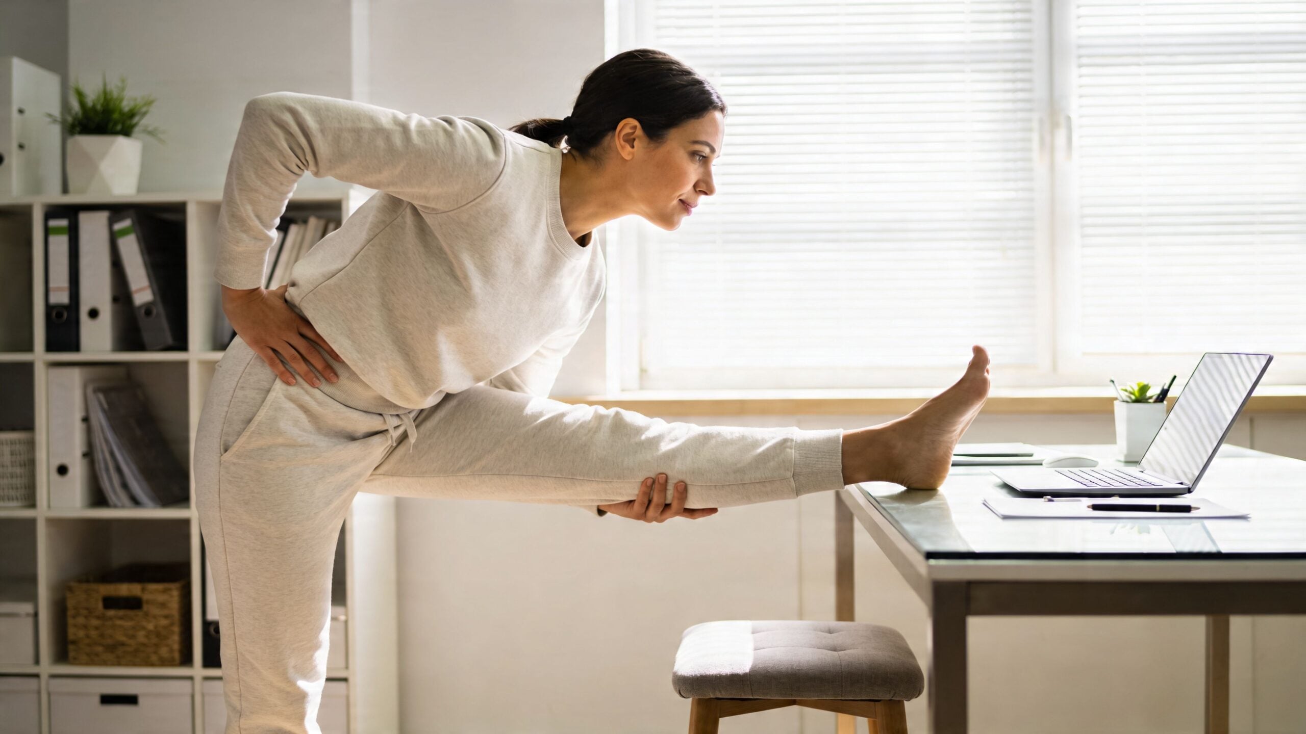 A young woman performing a standing hamstring stretch on a desk to relieve painful hips after sitting.