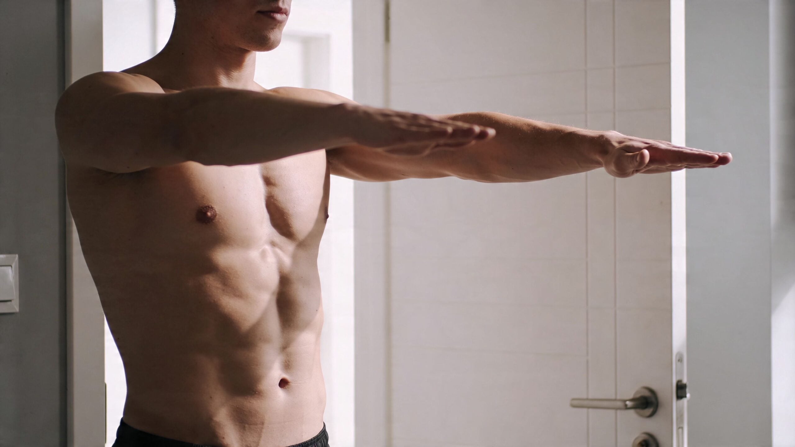 A shirtless muscular man performing a chest stretch by extending his arms forward in a doorway