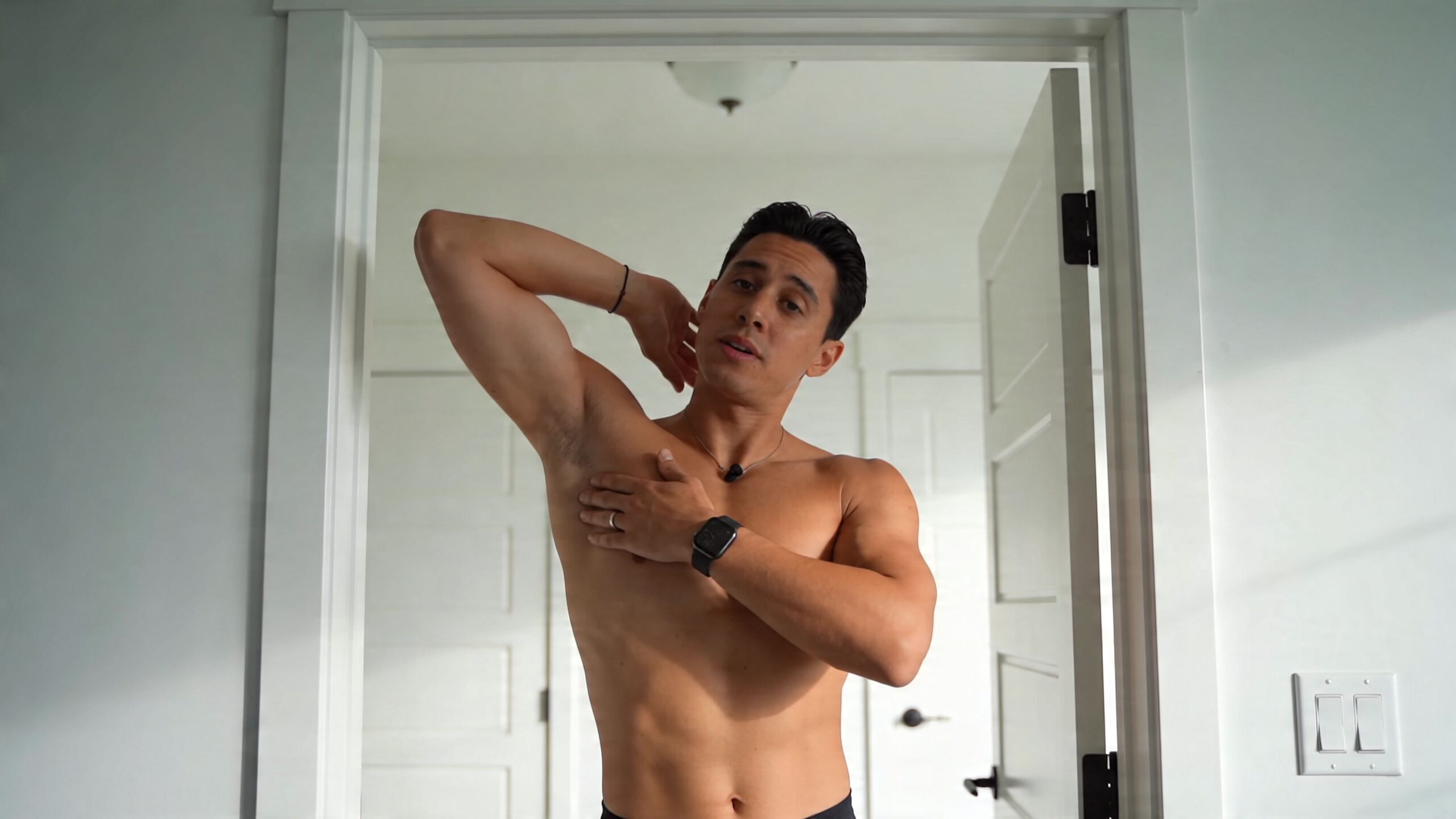 A fit shirtless man demonstrating a pectoral stretch by leaning against a doorway in a bright room.