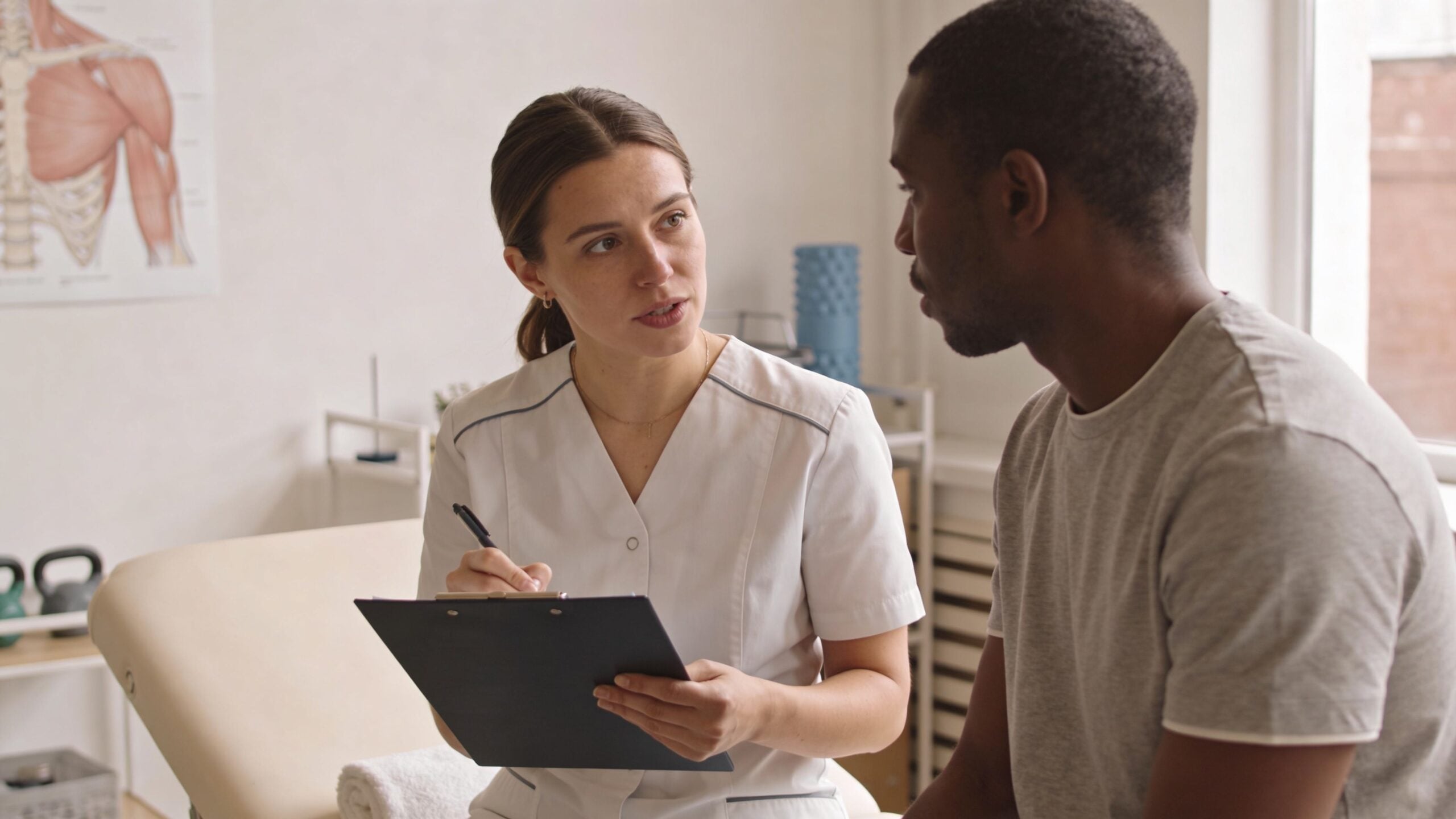 A physical therapist in a medical uniform consults with a patient while taking notes on a clipboard.