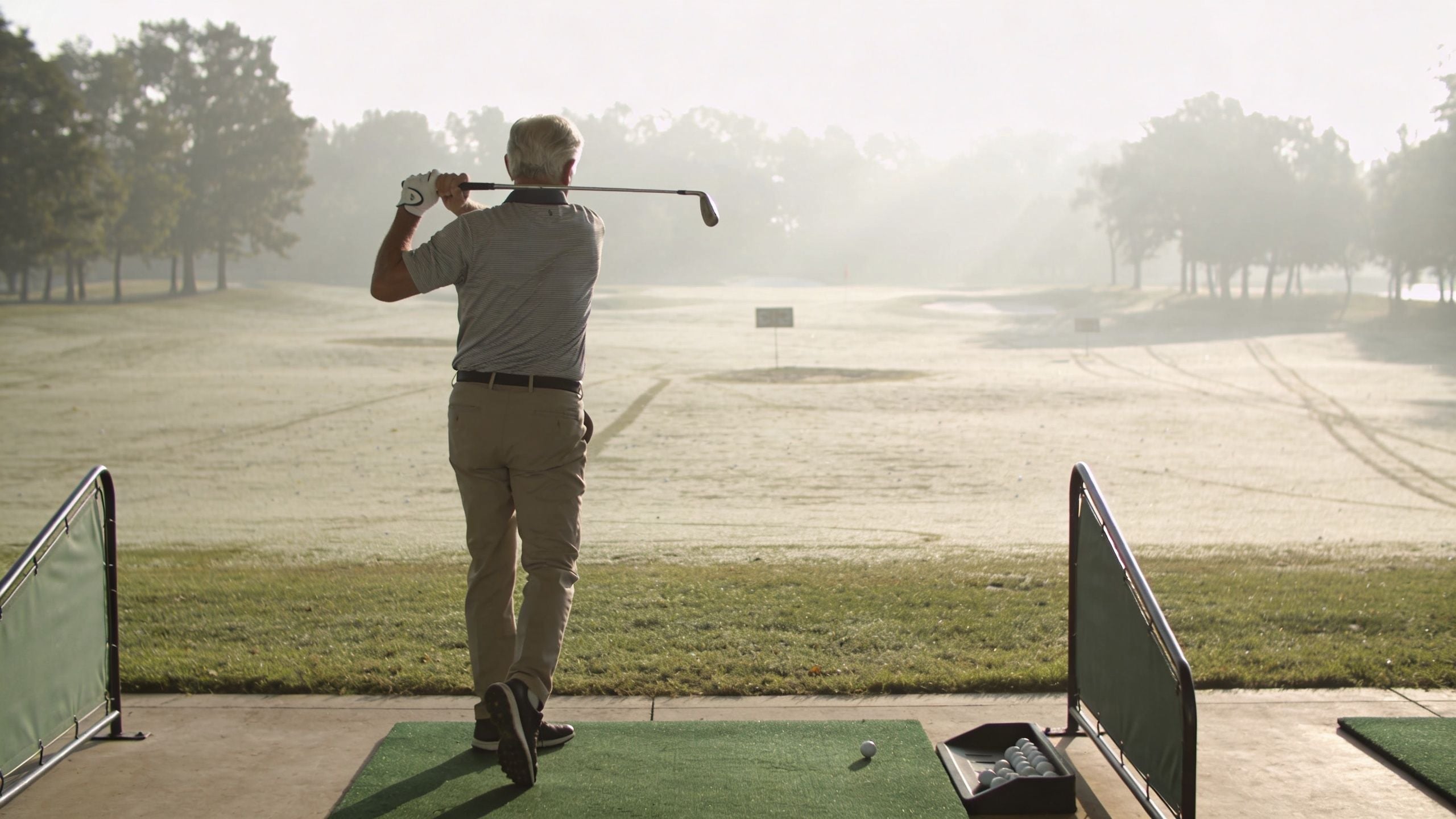 A senior man practicing his golf swing on a driving range during a foggy morning.