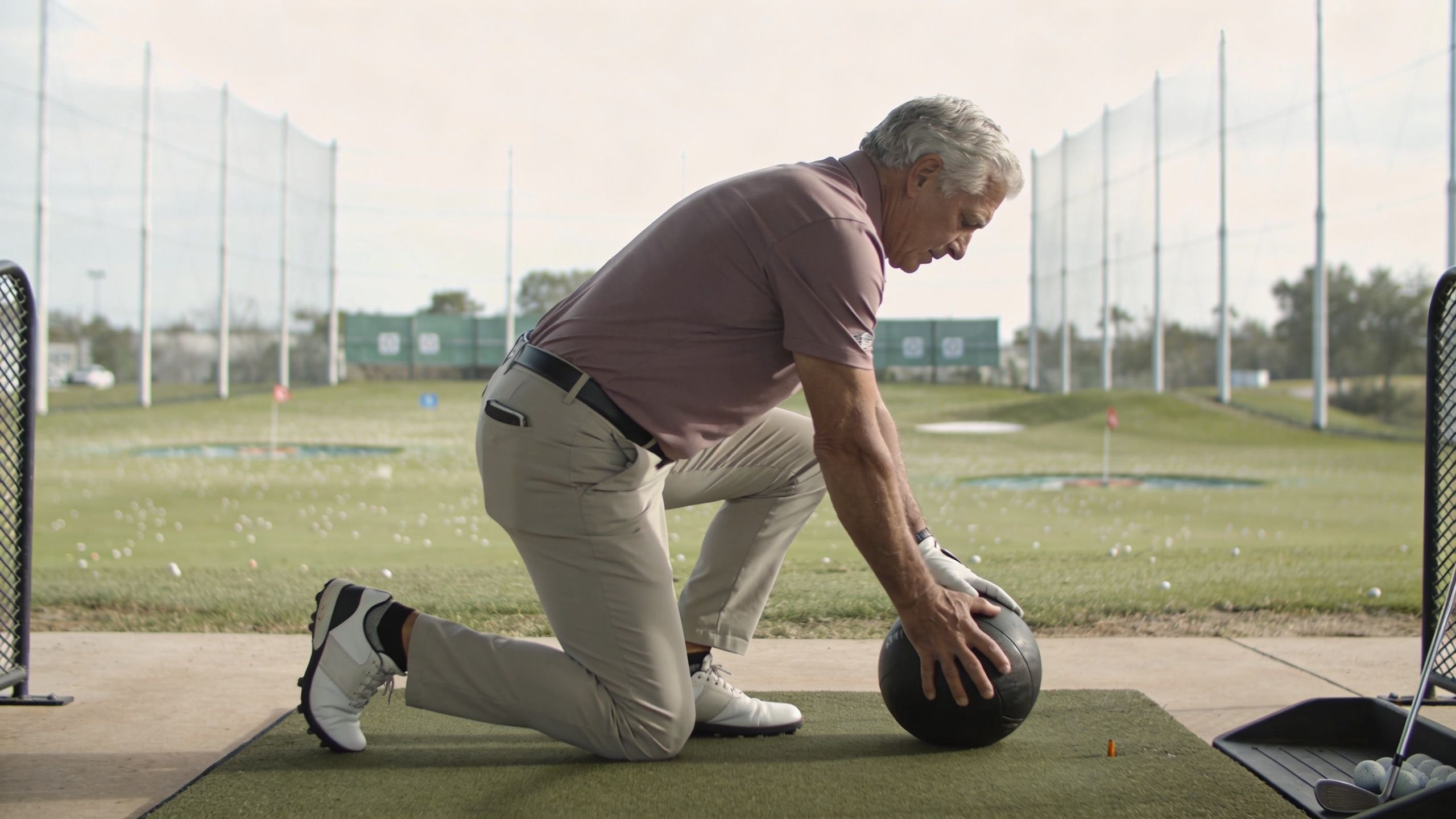 A senior golfer performing a stretching exercise with a medicine ball on a golf driving range mat.