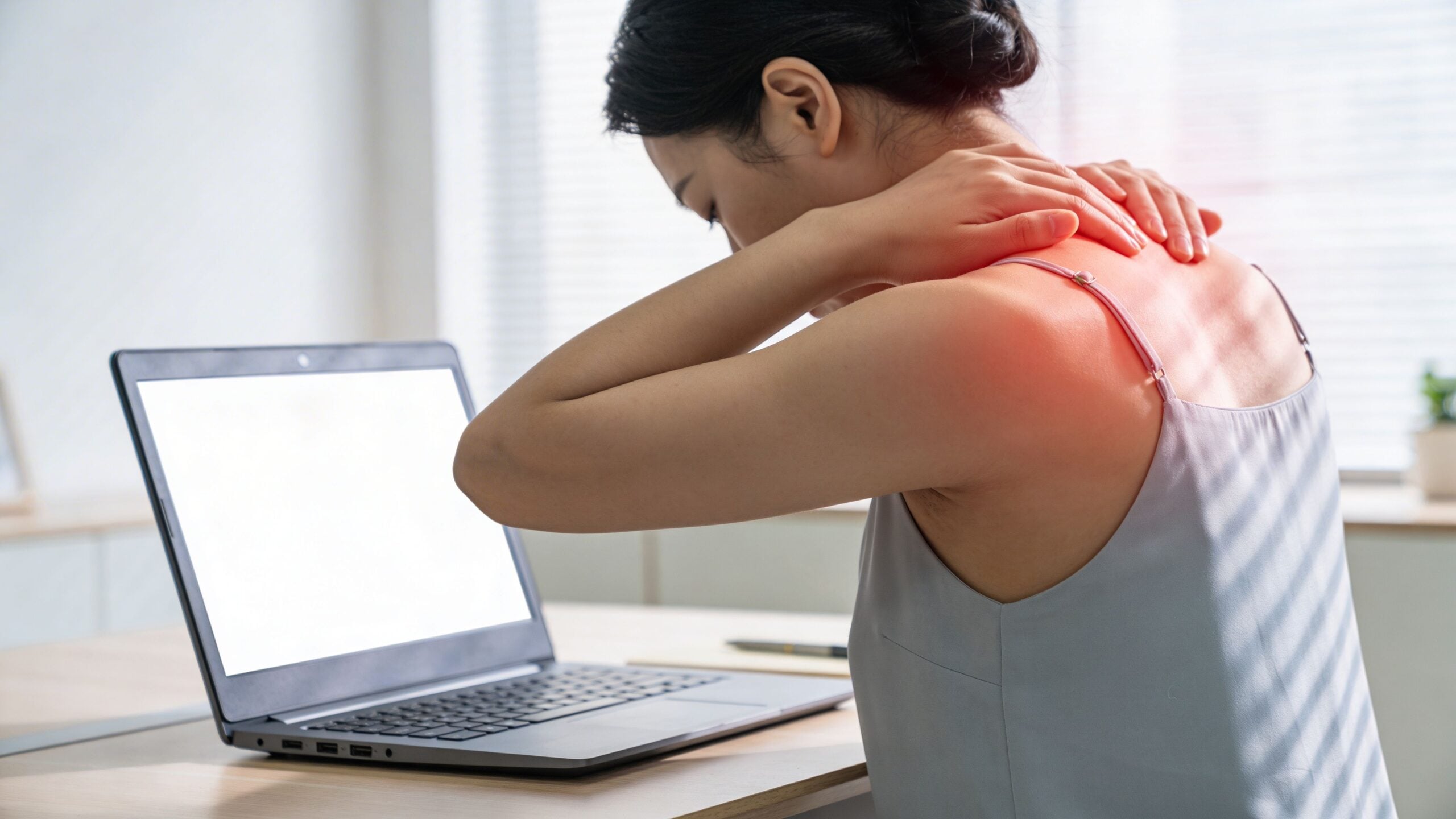 A woman experiencing neck and shoulder pain while working at a computer in an office setting.