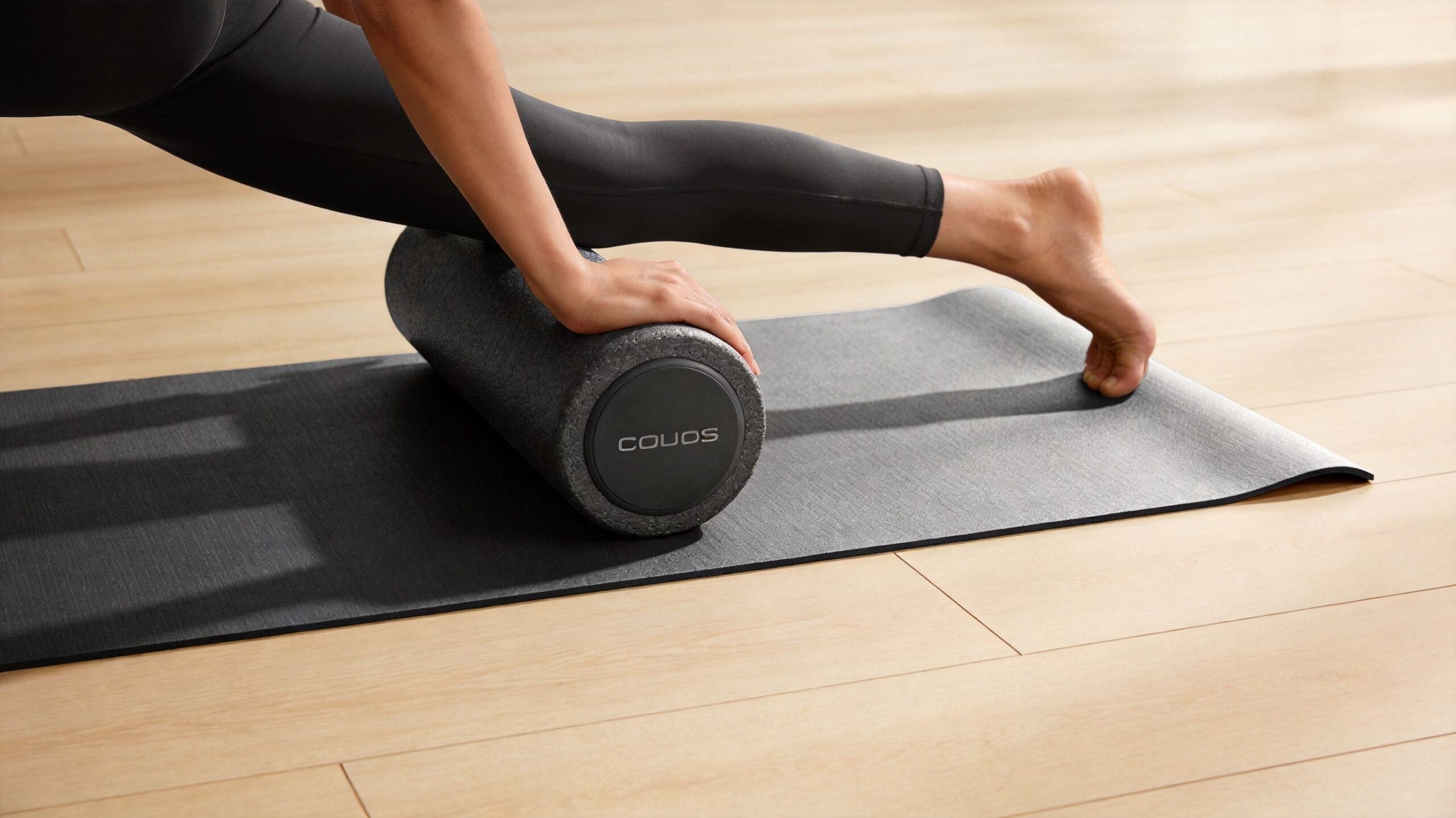 A person uses a grey foam roller on their leg while exercising on a black yoga mat.