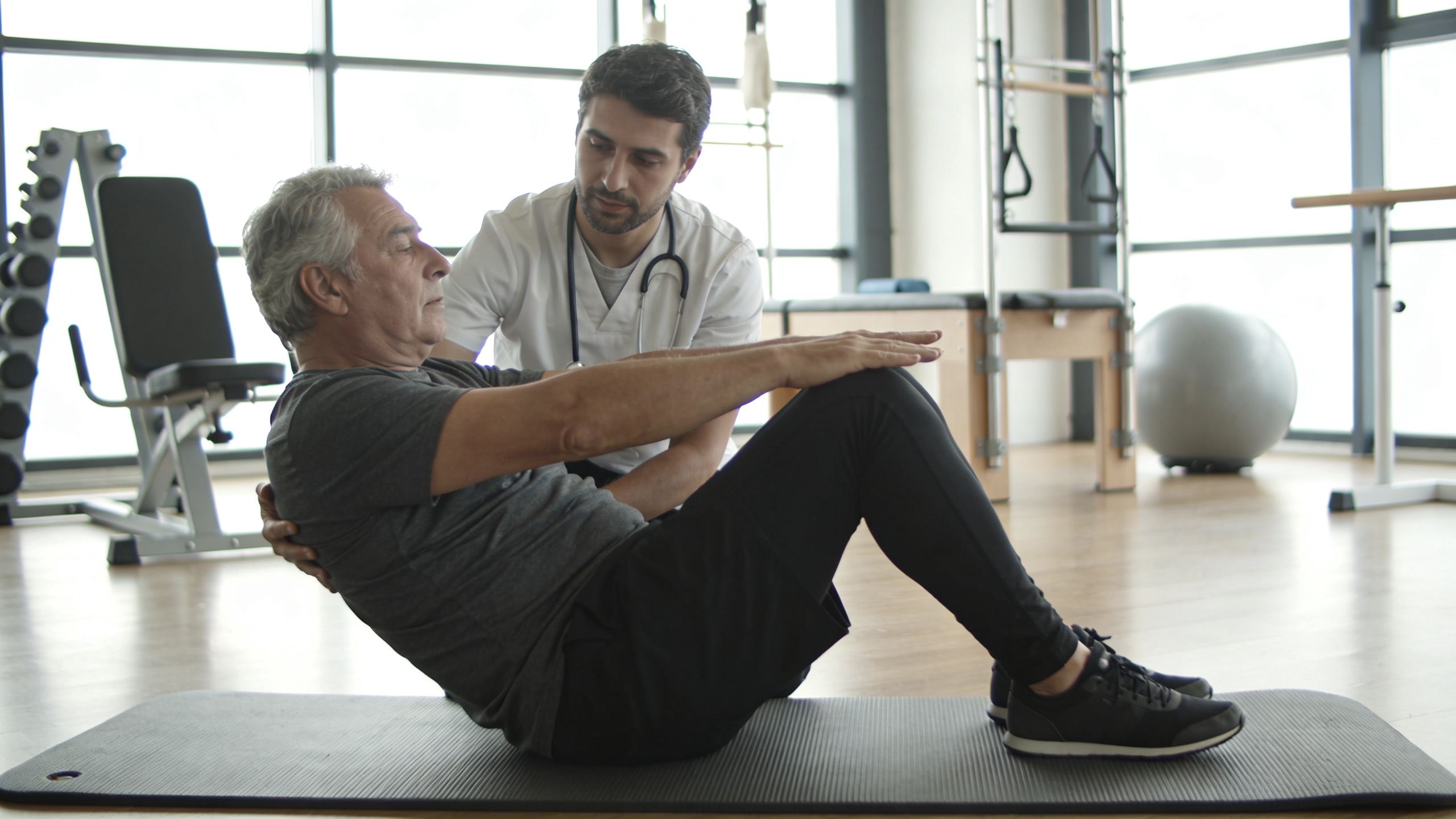 A male physical therapist assists an older man performing a sit-up exercise on a mat in a studio.